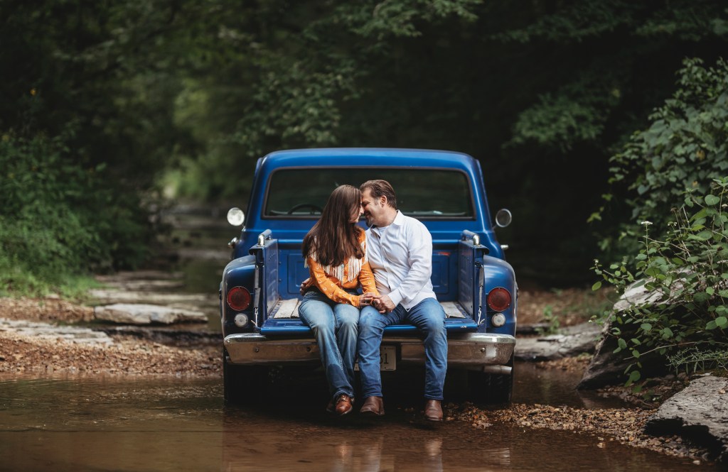 A couple sitting in the back of a blue pickup truck parked in a shallow creek surrounded by trees.