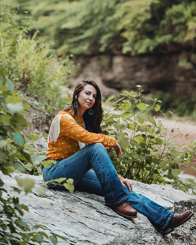 Woman relaxing on a rock near a creek in a natural setting, dressed casually in a yellow patterned blouse and jeans.